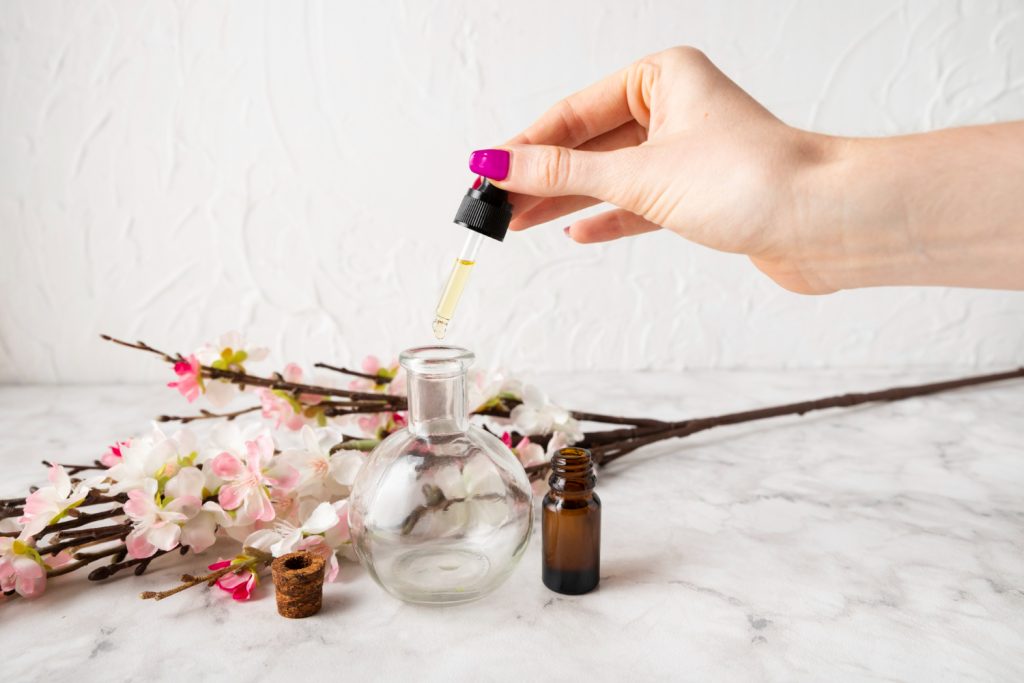 Indonesian Body Fragrance: A hand with bright nails drips oil into a glass bottle, surrounded by delicate pink blossoms and a dark amber vial.