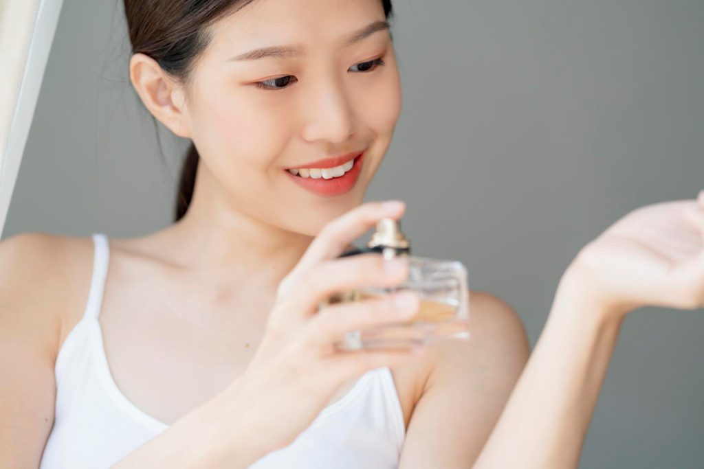 Body Fragrances shown by an Asian woman applies perfume from a bottle, showcasing elegance and self-care in a softly lit, neutral-toned setting.