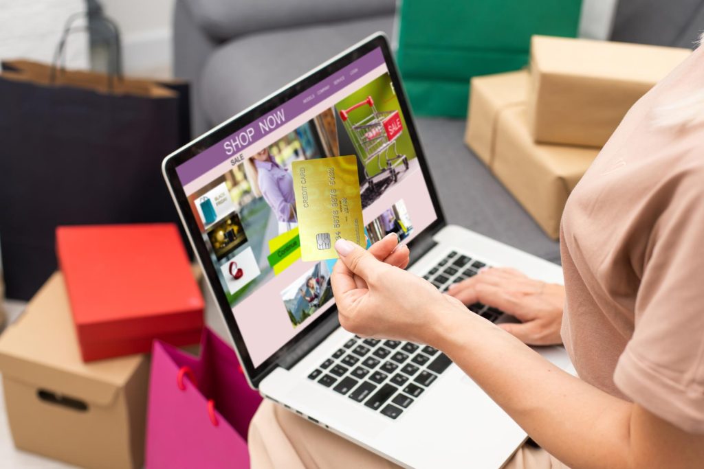 A woman shopping online holds a credit card while browsing a sale on her laptop, surrounded by shopping bags and packages.