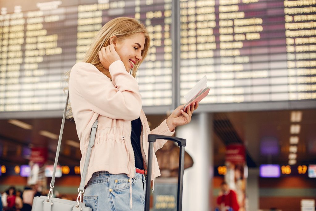 A traveler stands at an airport terminal, holding a ticket and looking at a flight information board, with a suitcase beside her.