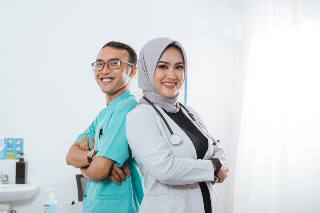 A male and female medical professional stand back-to-back in a bright clinic, arms crossed, showcasing a confident partnership.