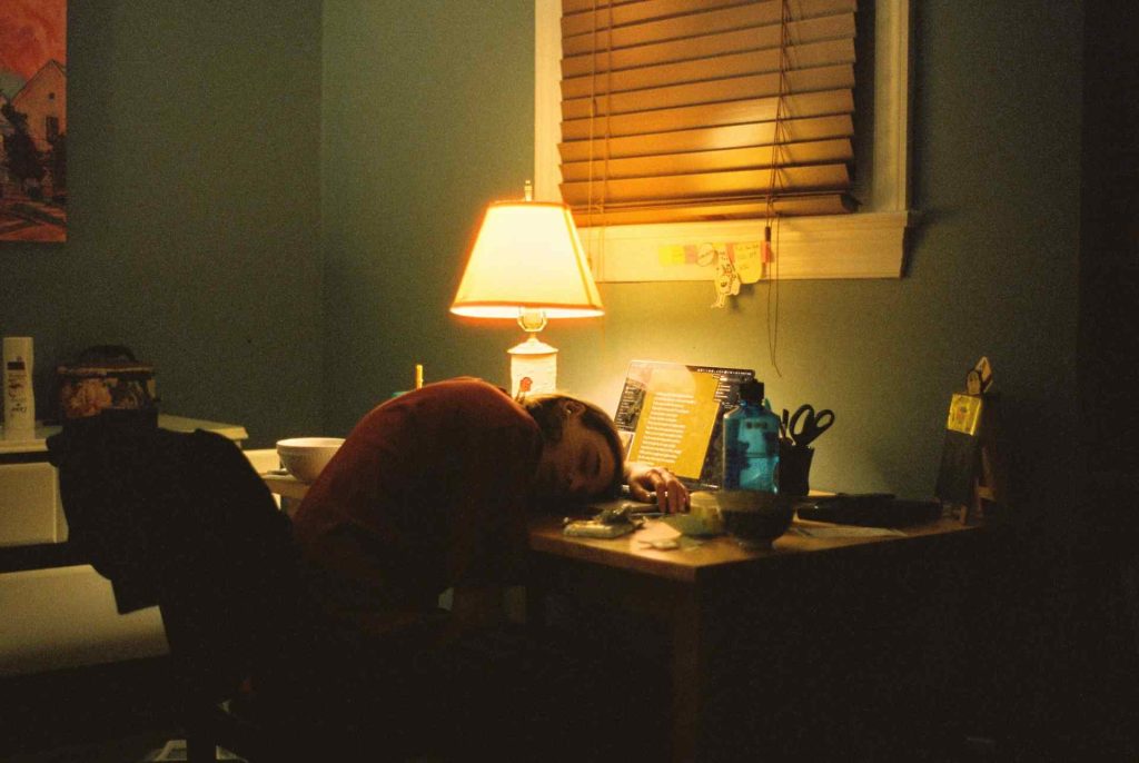A person leans their head on a desk in a dimly lit room, surrounded by scattered items, a glowing lamp, and a laptop.