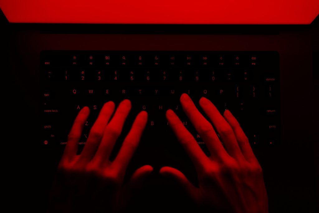 A close-up of hands typing on a backlit keyboard illuminated in red light, creating a dramatic and moody atmosphere.