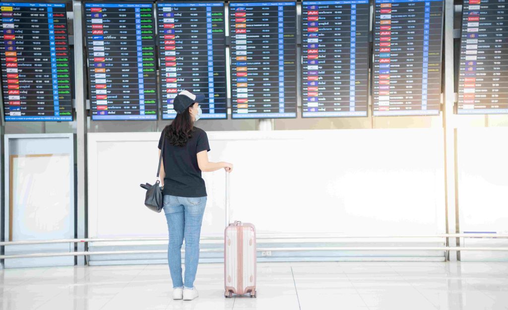 A woman with a suitcase stands gazing at flight information screens in an airport, wearing a black t-shirt, jeans, and a cap.