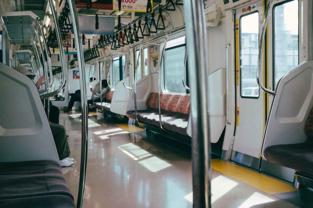 Interior of a subway train with empty seats, poles, and passengers reading or using their phones, illuminated by natural light.