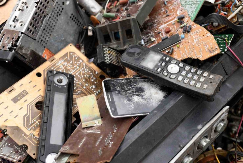 A pile of discarded electronic waste, including a cracked smartphone, old remote controls, and circuit boards.