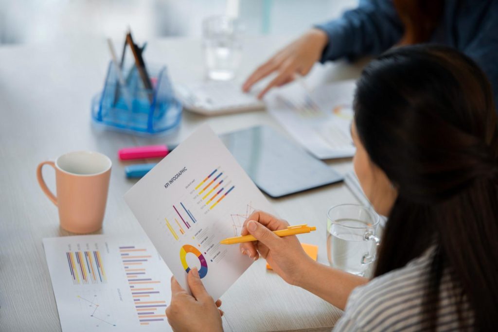 A person reviewing a colorful KPI infographic with graphs, holding a yellow pen, amidst a collaborative workspace with drinks and stationery.
