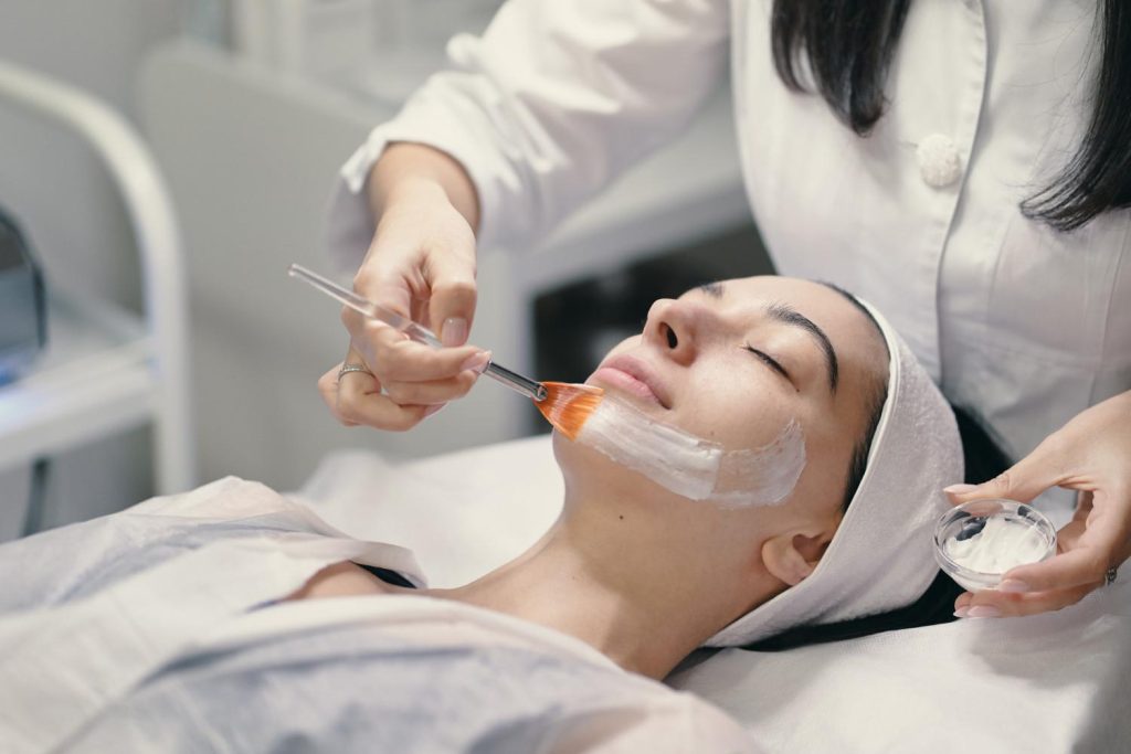 Indonesian beauty clinic market: A beautician applies a red facial mask using a brush on a client lying on a treatment bed, surrounded by skincare products.