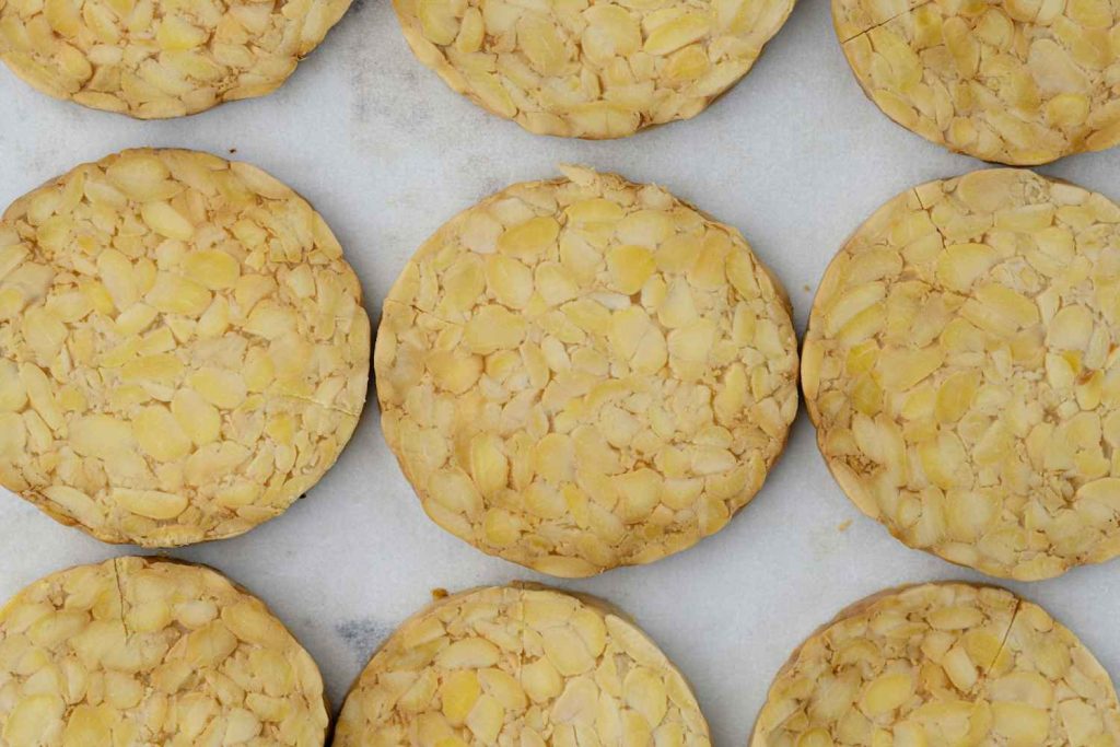A close-up of golden-brown tempeh, flat and round, scattered on a white baking paper.