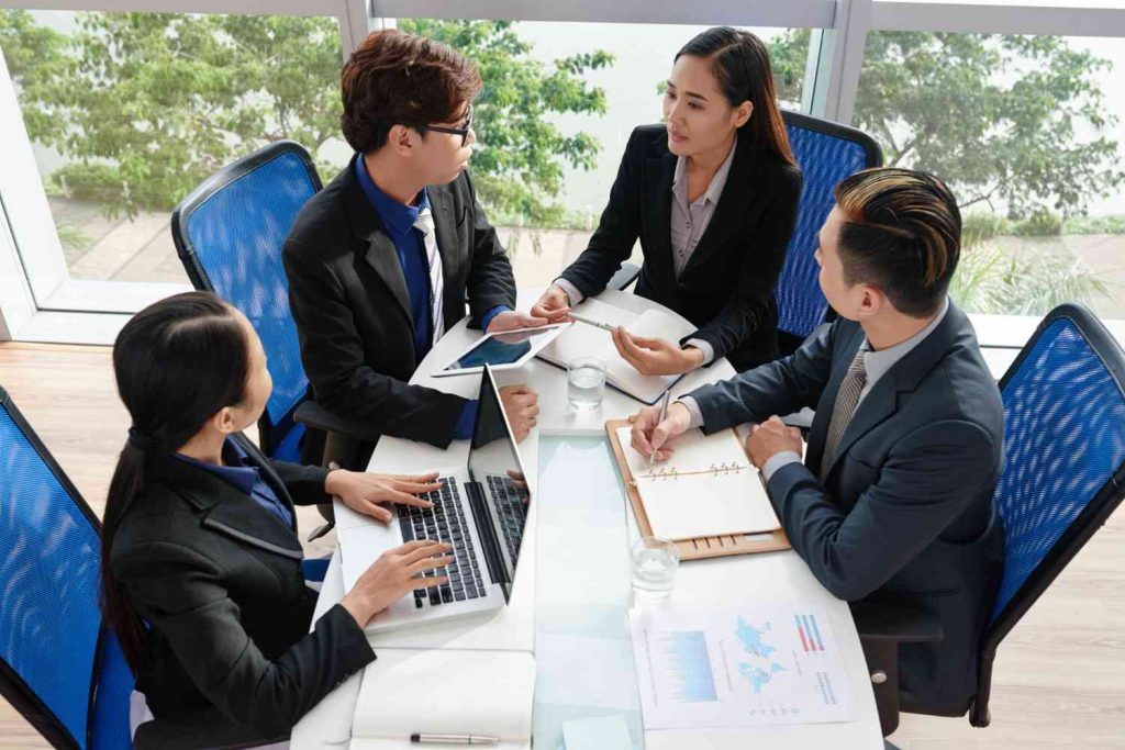 A group of four professionals in suits engage in a meeting around a glass table, with laptops, tablets, and documents displayed.