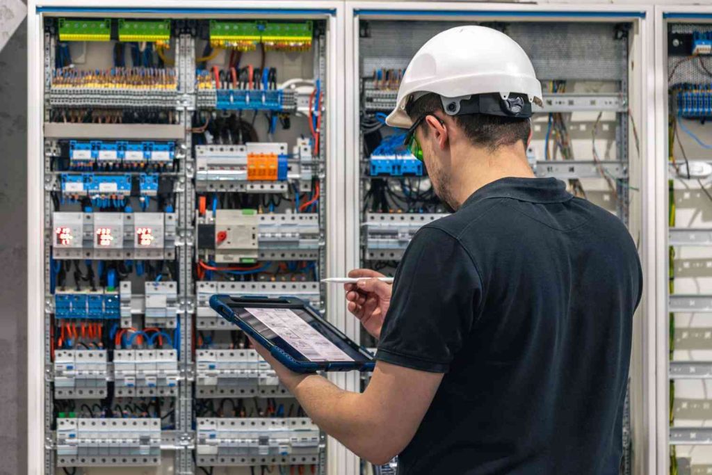 A worker in a safety helmet inspects electrical panels while using a tablet, surrounded by various wiring and components.