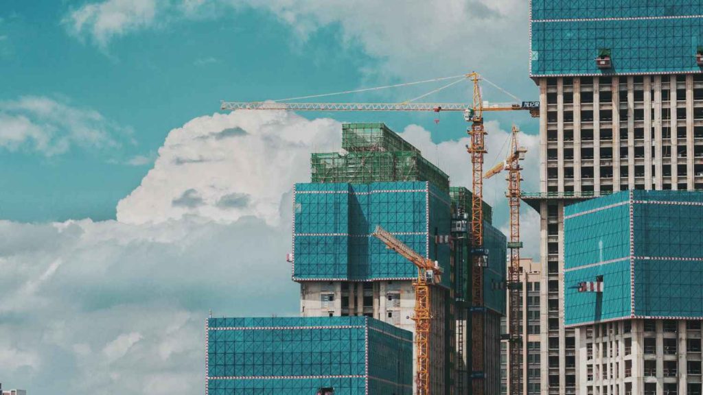Construction site of a high-rise building with cranes, blue scaffolding, and cloudy skies in the background.